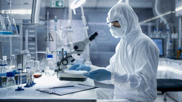 A scientist dressed in full protective gear, including a white suit, gloves, and goggles, is working in a laboratory. They are using a microscope on a desk surrounded by various lab equipment, such as test tubes, beakers, and bottles.