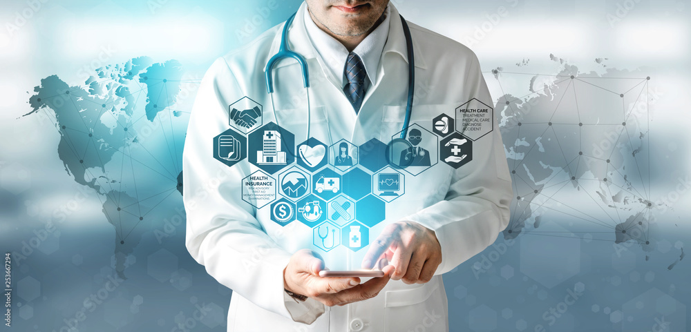 A doctor in a white lab coat with a stethoscope around his neck uses a tablet. Transparent medical icons, such as a heart, pill, and hospital, are projected above the tablet. A blurred world map is in the background, symbolizing global healthcare connectivity.