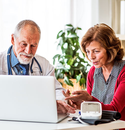 A doctor with gray hair and a stethoscope, seated beside a middle-aged woman, discussing something displayed on a laptop screen. A blood pressure monitor is placed on the table in front of them, and there is a plant in the background.