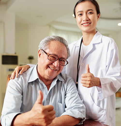 An elderly man wearing glasses and a light blue shirt gives a thumbs-up while sitting. Beside him stands a smiling woman in a white coat, also showing a thumbs-up gesture. They are indoors, in a well-lit room with neutral colors.