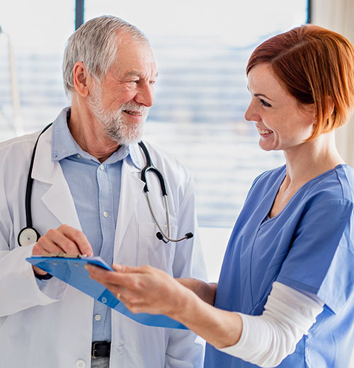 A smiling older male doctor with grey hair and beard in a white coat and stethoscope discusses information on a clipboard with a smiling younger female nurse with red hair in blue scrubs. They are indoors with a window in the background.