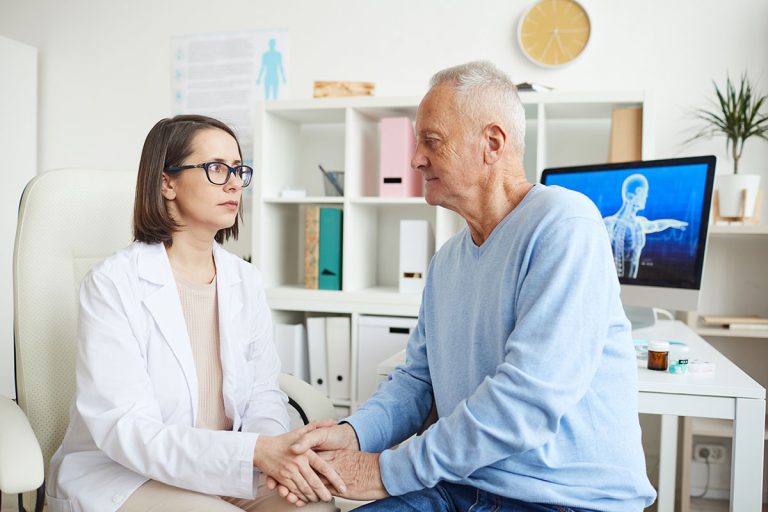 A doctor and an elderly patient are in a medical office. The doctor, wearing glasses and a white coat, is holding the patient's hand and looking at him attentively. The patient, dressed in a light blue sweater, is seated and gazing back at the doctor. A computer with medical imagery is in the background.