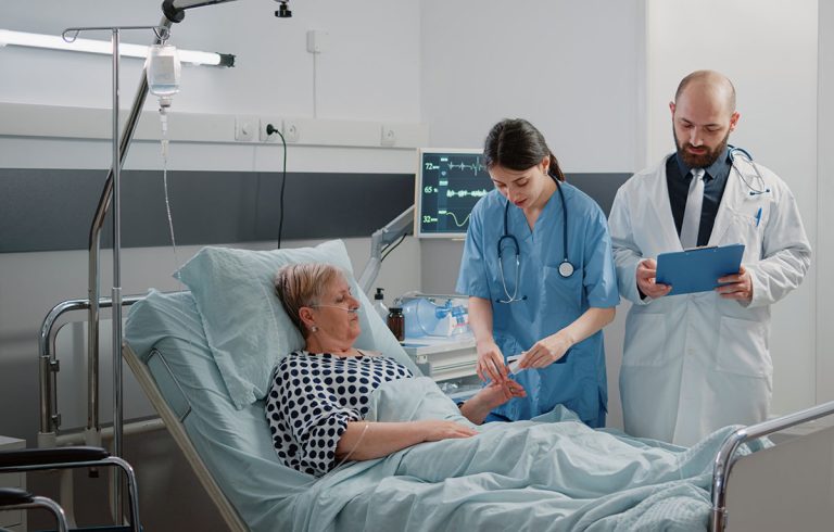 A patient lying in a hospital bed receives care from two medical professionals. One is a nurse adjusting an IV drip, while the other is a doctor holding a clipboard. Medical equipment and monitors surround the patient's bed in the hospital room.